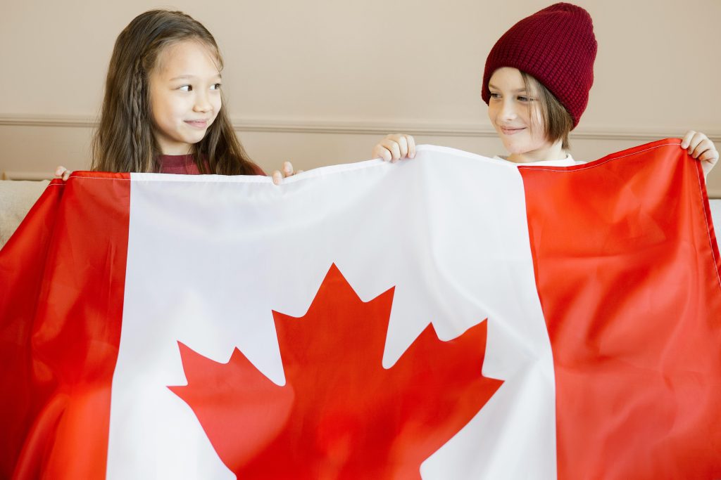 Two children joyfully holding the Canadian flag indoors, celebrating national pride.